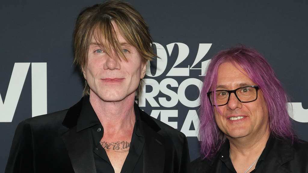 Goo Goo Dolls members John Rzeznik, with light brown hair and a slight smile, and Robby Takac, with purple hair and glasses, pose at an event.