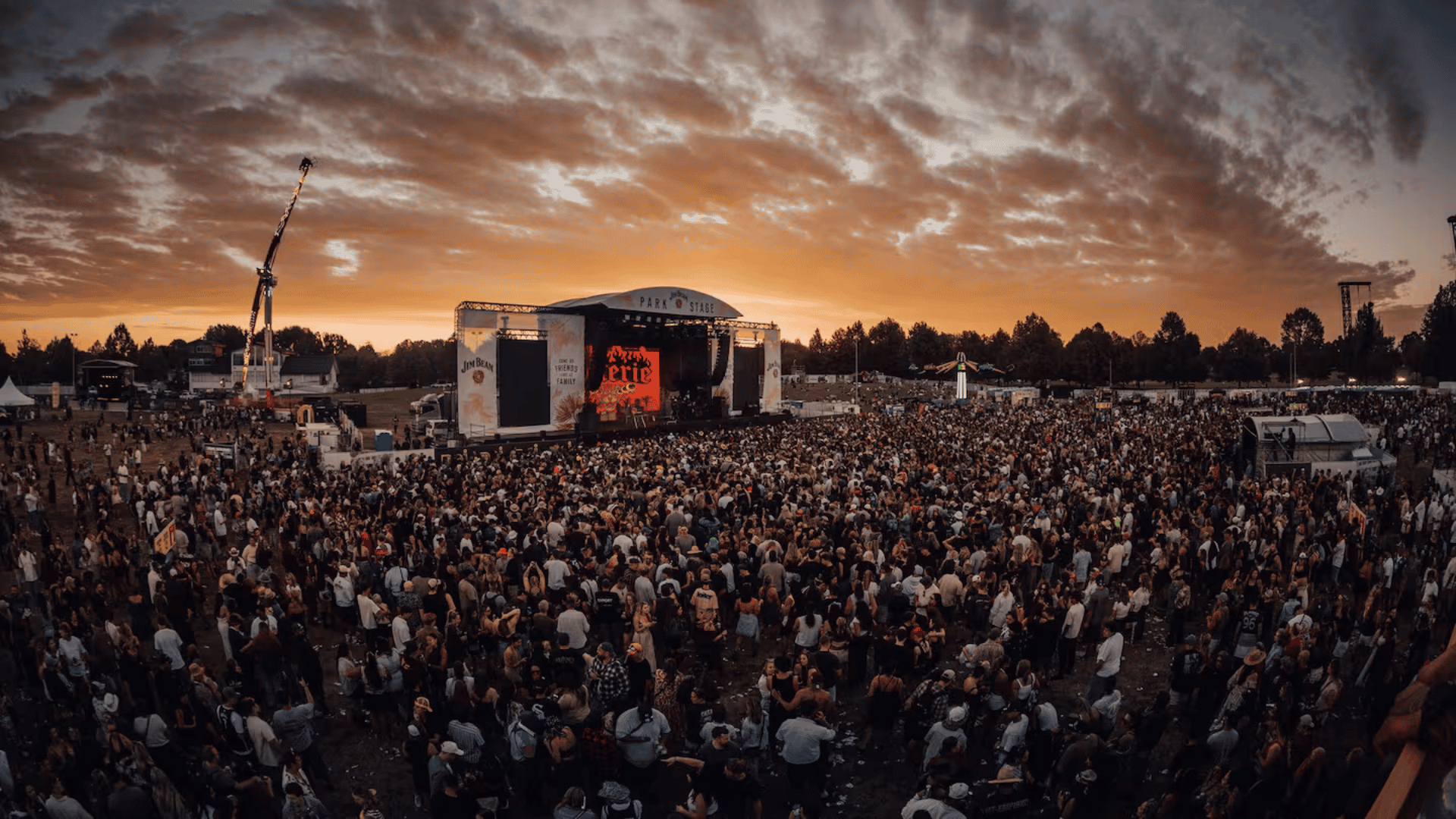 A wide shot of a large outdoor music festival with a massive crowd in front of the "PARK STAGE" under a cloudy orange sunset sky, featuring Jim Beam branding.