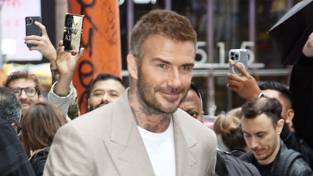 David Beckham, with a short beard and visible neck tattoos, smiles at the camera, wearing a light suit jacket and white t-shirt, surrounded by people holding up phones.