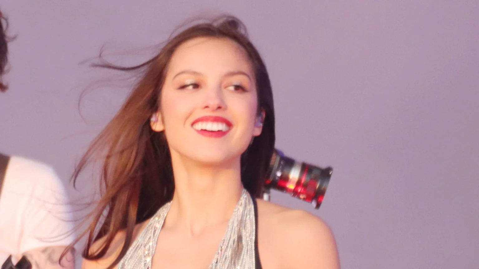 A young woman with brown hair blowing in the wind smiles widely, showing teeth, with red lipstick and a sparkly silver top, against a plain purple background.