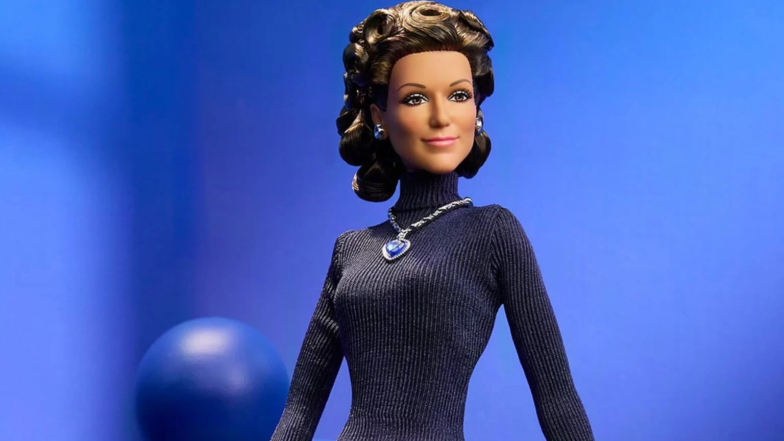 A Barbie doll with dark, curly hair and brown eyes wears a dark blue ribbed turtleneck, silver earrings, and a large heart-shaped blue gem necklace against a blue background.