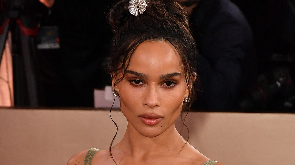 Zoë Kravitz looks directly at the camera, wearing a high messy bun with a silver hairpiece, small earrings, and a light green dress.
