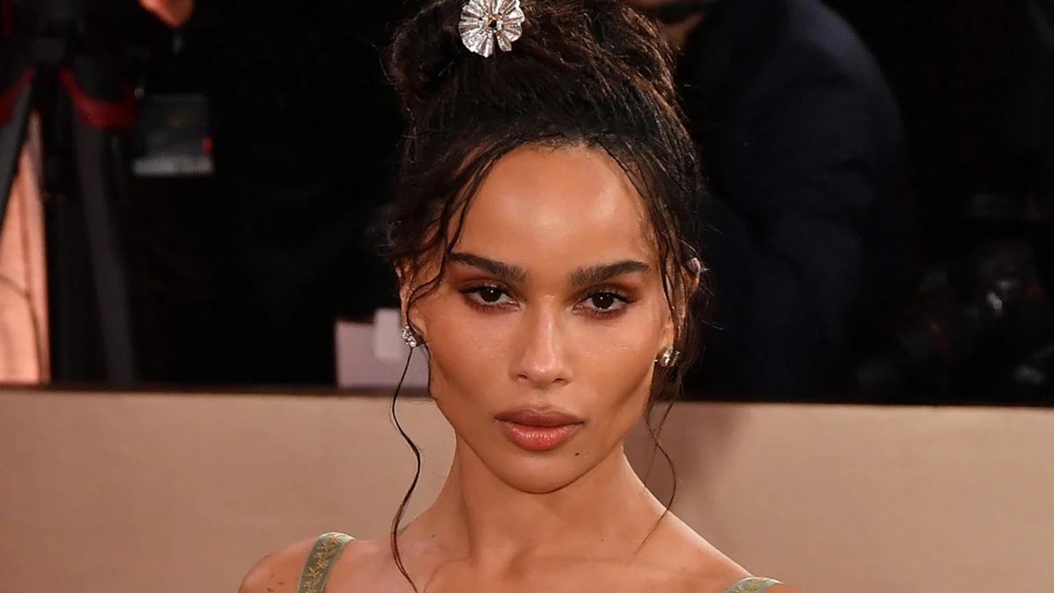 Zoë Kravitz looks directly at the camera, wearing a high messy bun with a silver hairpiece, small earrings, and a light green dress.
