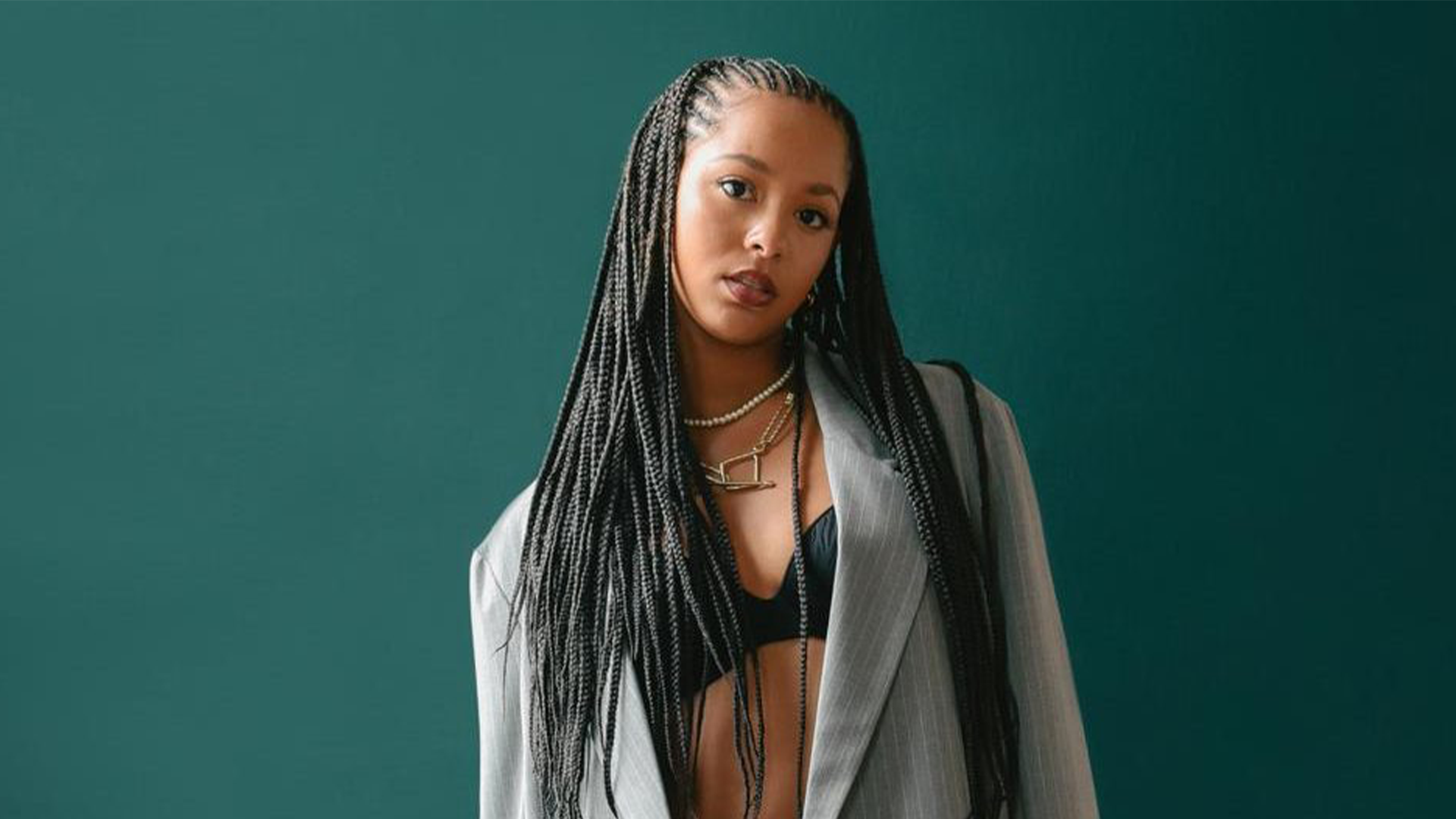 A young woman with long braids looks at the camera, wearing a black bralette, grey pinstripe blazer, and layered necklaces against a teal background.