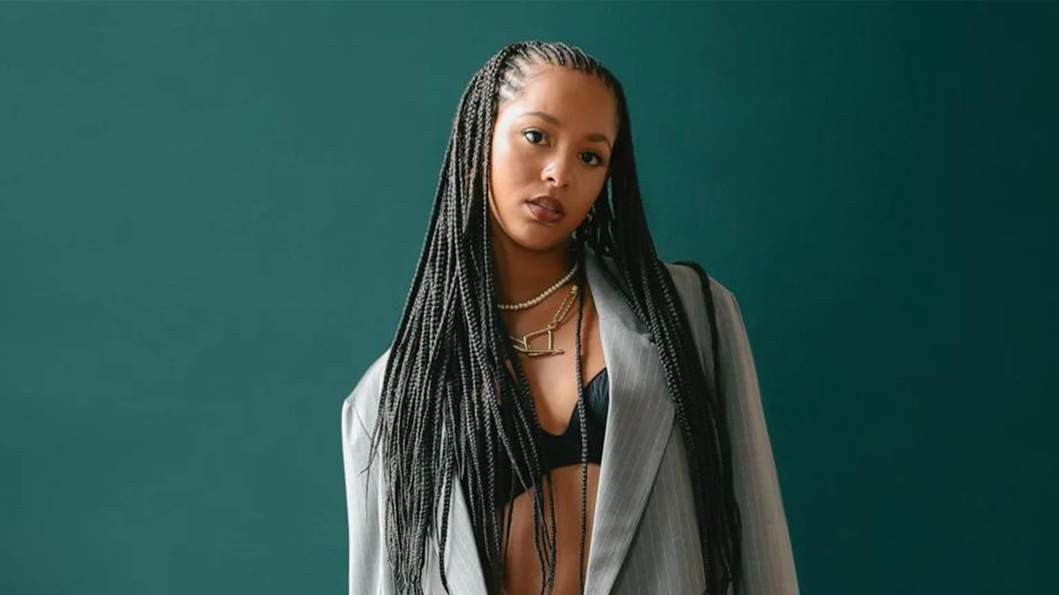 A young woman with long braids looks at the camera, wearing a black bralette, grey pinstripe blazer, and layered necklaces against a teal background.