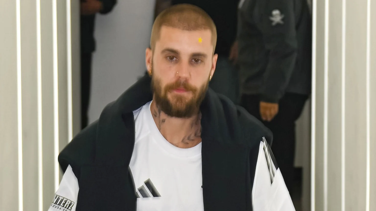 A man with a short beard and hair, wearing a white Adidas shirt and a dark jacket, looks at the camera. He has a yellow star sticker on his forehead and visible neck tattoos.