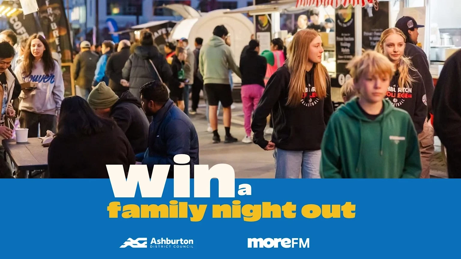 People gather and walk at a lively nighttime food festival with food stalls visible in the background, with the prominent text "Win a family night out" at the bottom.