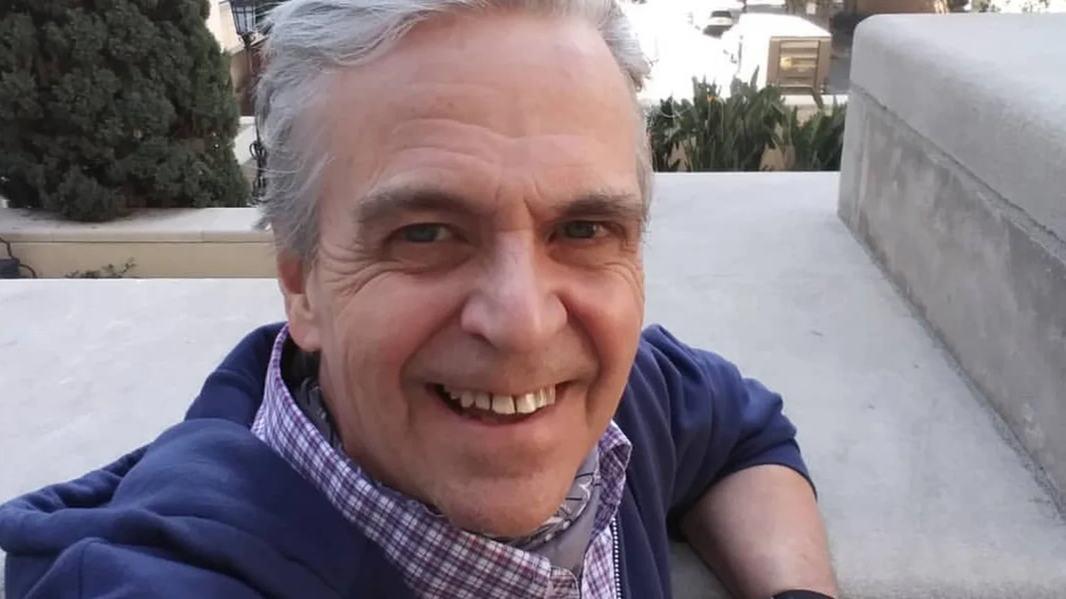 A smiling man with gray hair, blue eyes, and a gap between his front teeth wears a blue hoodie and a plaid shirt, looking directly at the camera.
