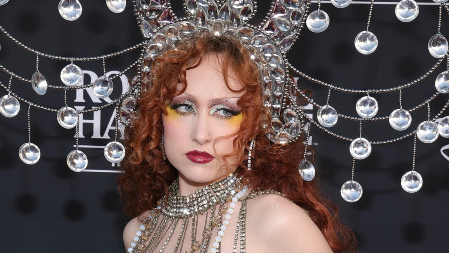 A woman with red curly hair and elaborate makeup, wearing a large jeweled crown with dangling silver discs and a sparkling necklace, looks to the side.