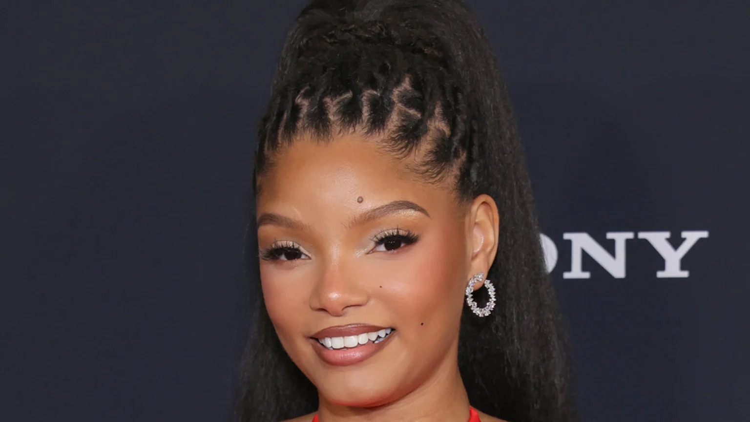 Close-up of Halle Bailey smiling, wearing a high ponytail with braids, sparkling hoop earrings, and natural makeup, against a dark background with "SONY" visible.