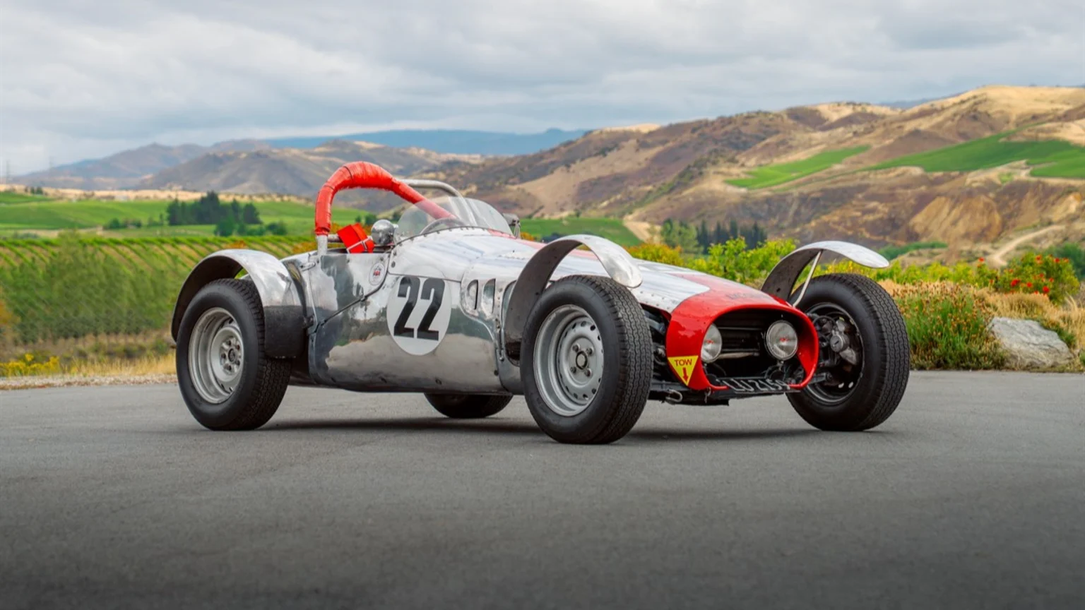A polished vintage-style open-wheel race car featuring a silver body with red accents and racing number 22 is parked on a smooth dark asphalt road. The backdrop showcases rolling hills with green vineyards, dry vegetation, flowering plants, and an overcast cloudy sky. A small yellow triangular 'TOW' sign and a red roll bar behind the driver are visible on the car.