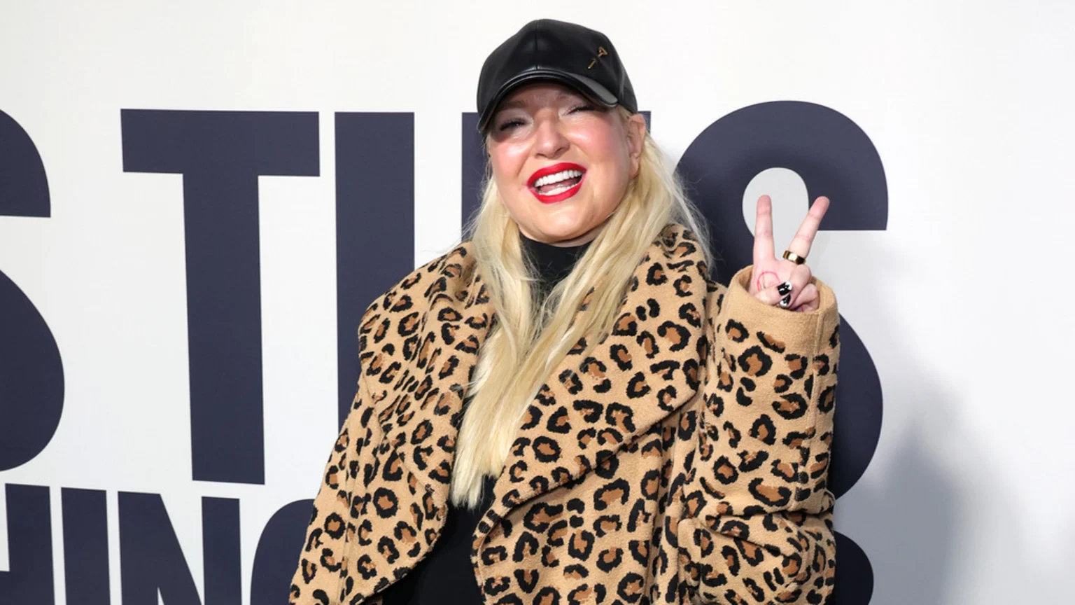 A blonde woman wearing a black cap, red lipstick, and a leopard print coat smiles and makes a peace sign. She stands before a white wall with dark blue letters.