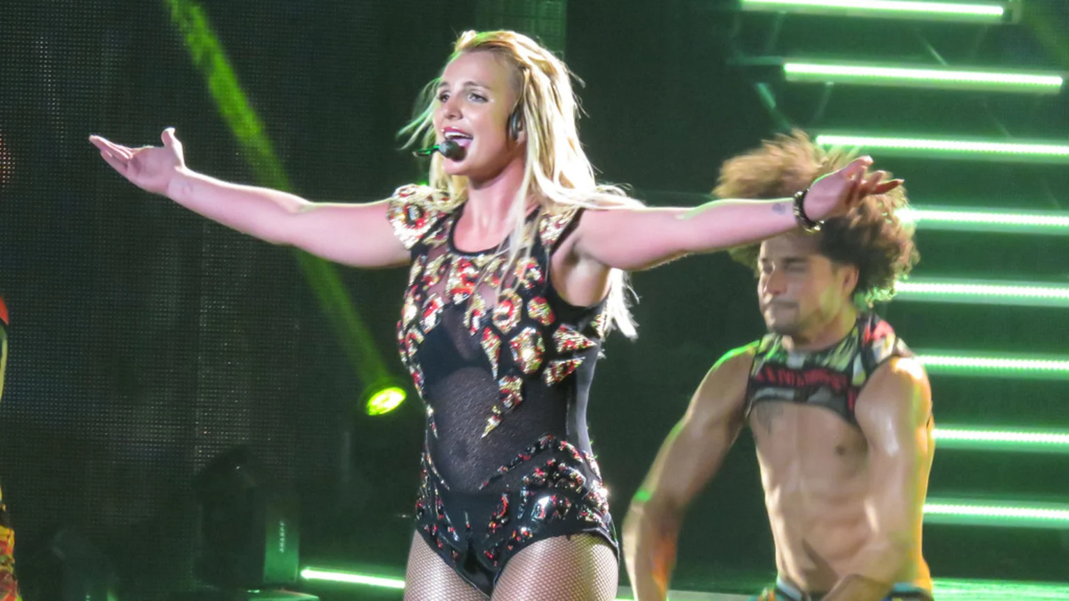 Britney Spears performs on stage in a sequined black bodysuit with outstretched arms, a male dancer behind her, and green lights in the background.