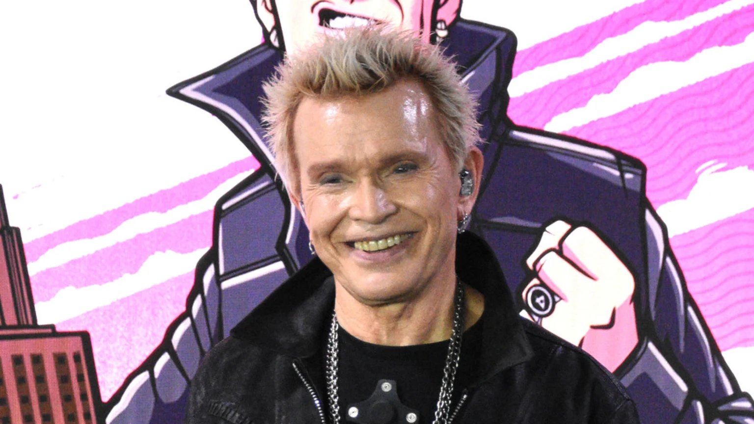 Smiling Billy Idol, with spiky blonde hair, wears a black leather jacket, chain necklaces, earrings, and in-ear monitors, against a vibrant cartoon-style background featuring a figure and a cityscape.