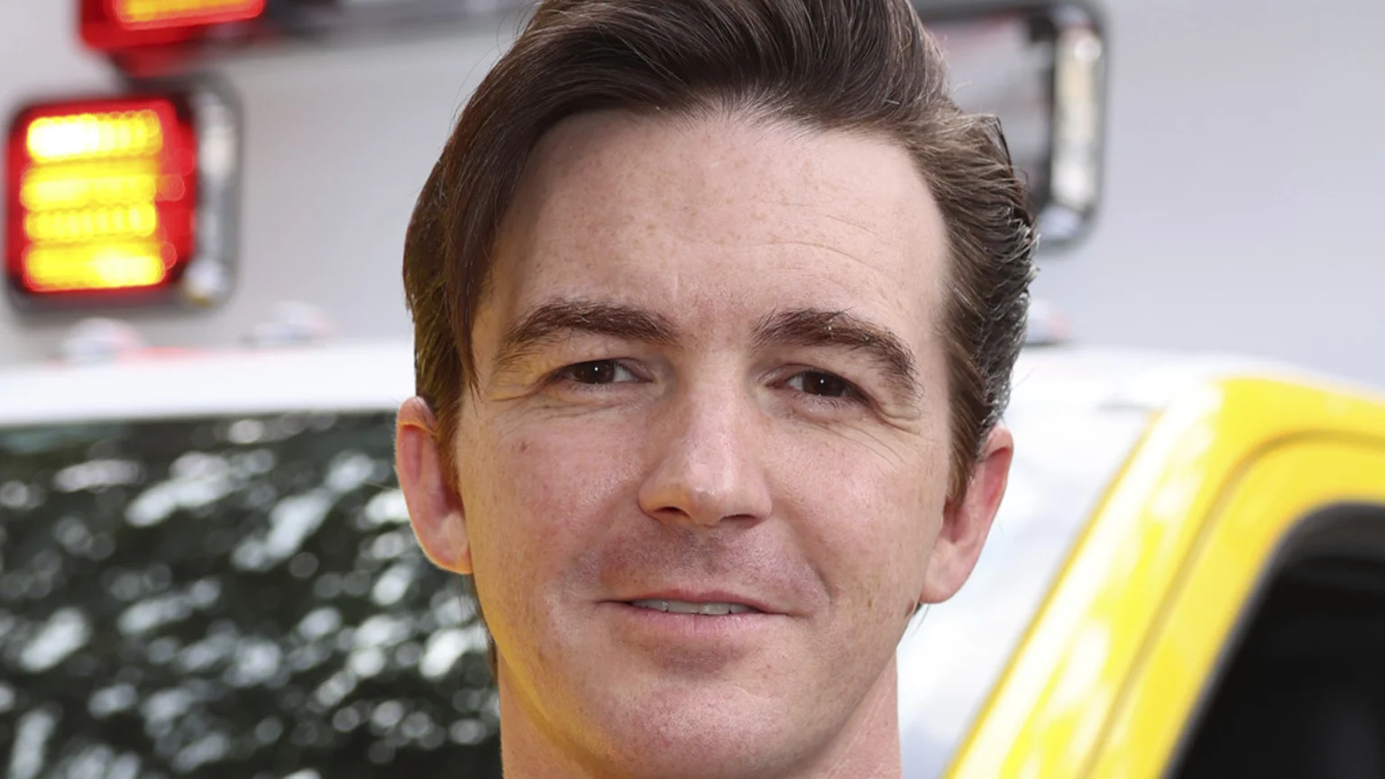 A close-up headshot of actor Drake Bell smiling faintly, with a blurred background showing red lights and part of a yellow vehicle.