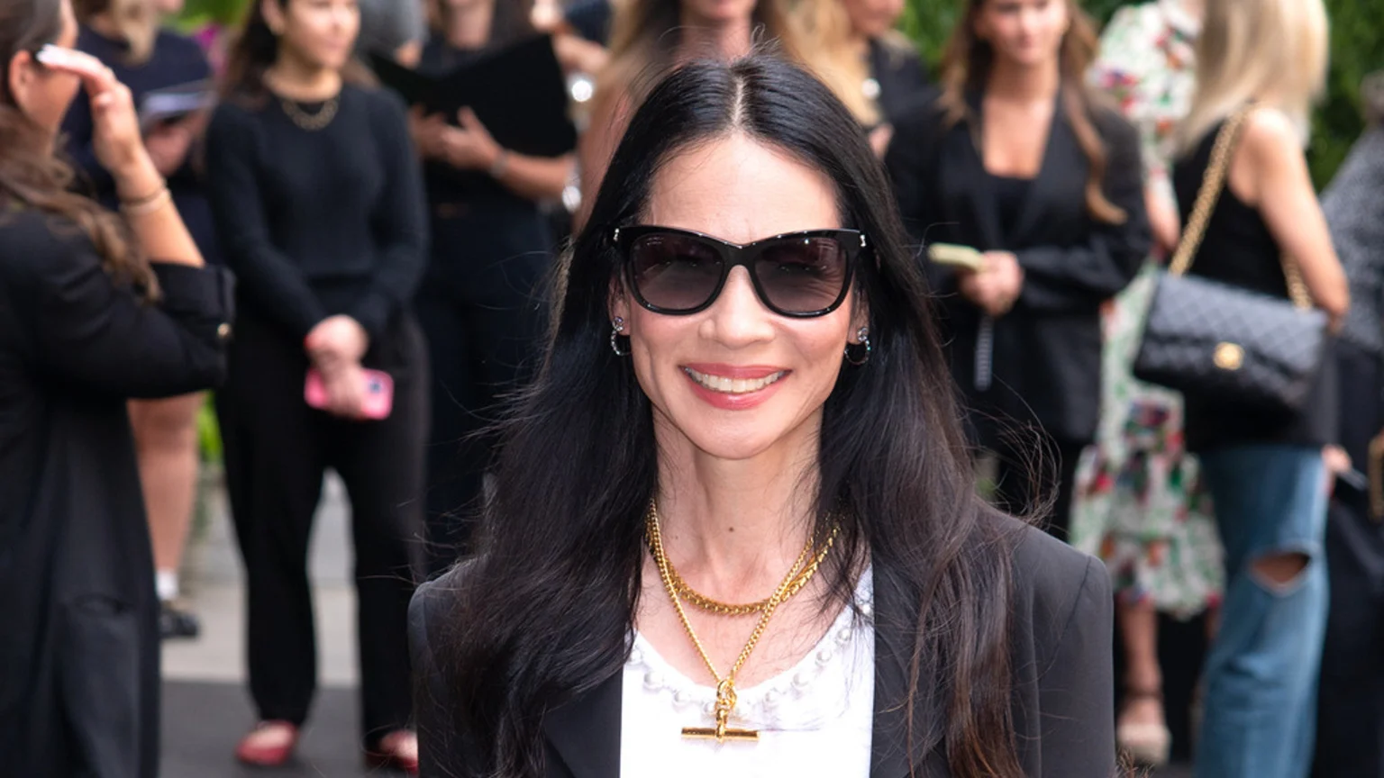 Lucy Liu smiles at the camera, wearing sunglasses, a black blazer over a white top, and layered gold necklaces, with blurry figures in the background.