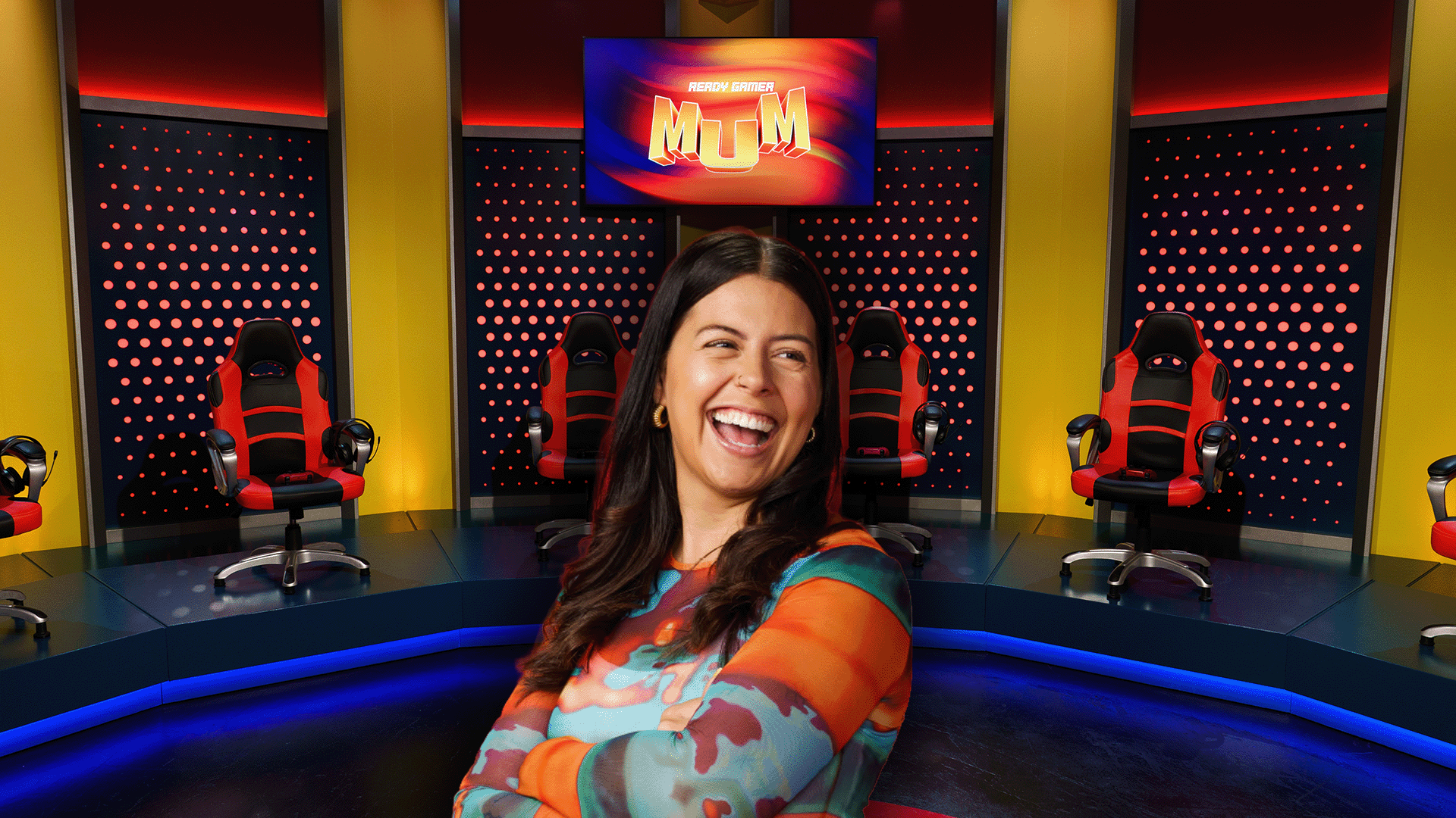 A laughing woman in a colorful top stands in front of a gaming show set with several red and black chairs and a screen displaying "READY GAMER MUM."