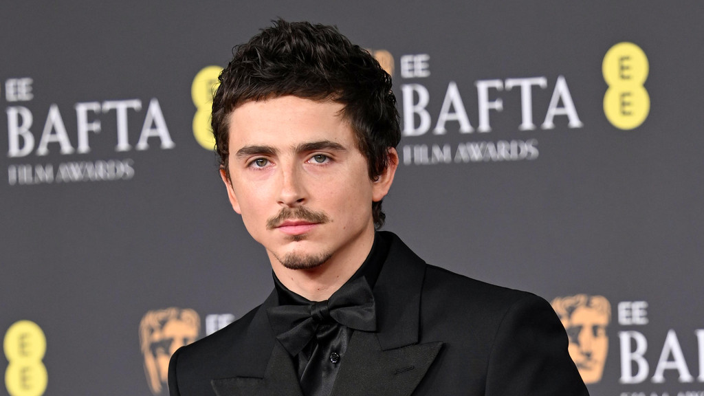 Timothée Chalamet, with a mustache and goatee, wearing a black suit, shirt, and bow tie, poses in front of an EE BAFTA Film Awards banner.