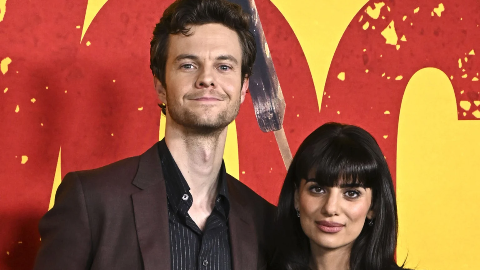 Jack Quaid and Claudia Domit pose together, smiling, against a red and yellow patterned background.