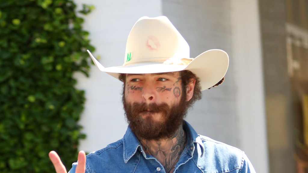 Post Malone, with face tattoos and a beard, wears a denim shirt and a light-colored cowboy hat with a pink lipstick print, making a peace sign.