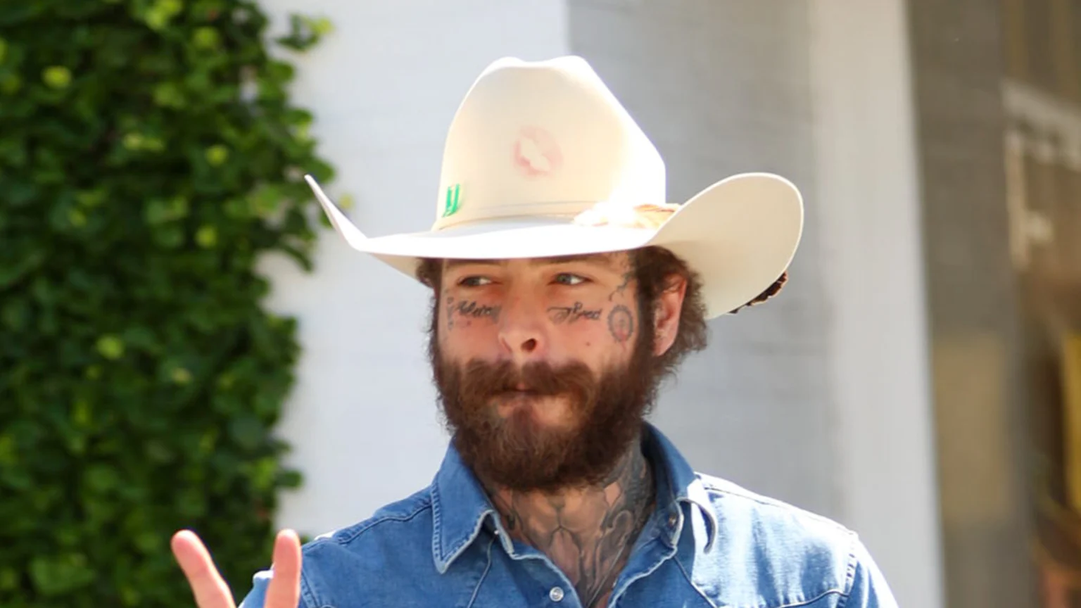 Post Malone, with face tattoos and a beard, wears a denim shirt and a light-colored cowboy hat with a pink lipstick print, making a peace sign.