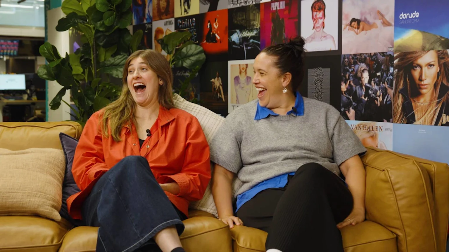 Two women on a light brown couch laugh heartily, one in an orange shirt, the other in a grey sweater, with a wall of various album covers behind them.
