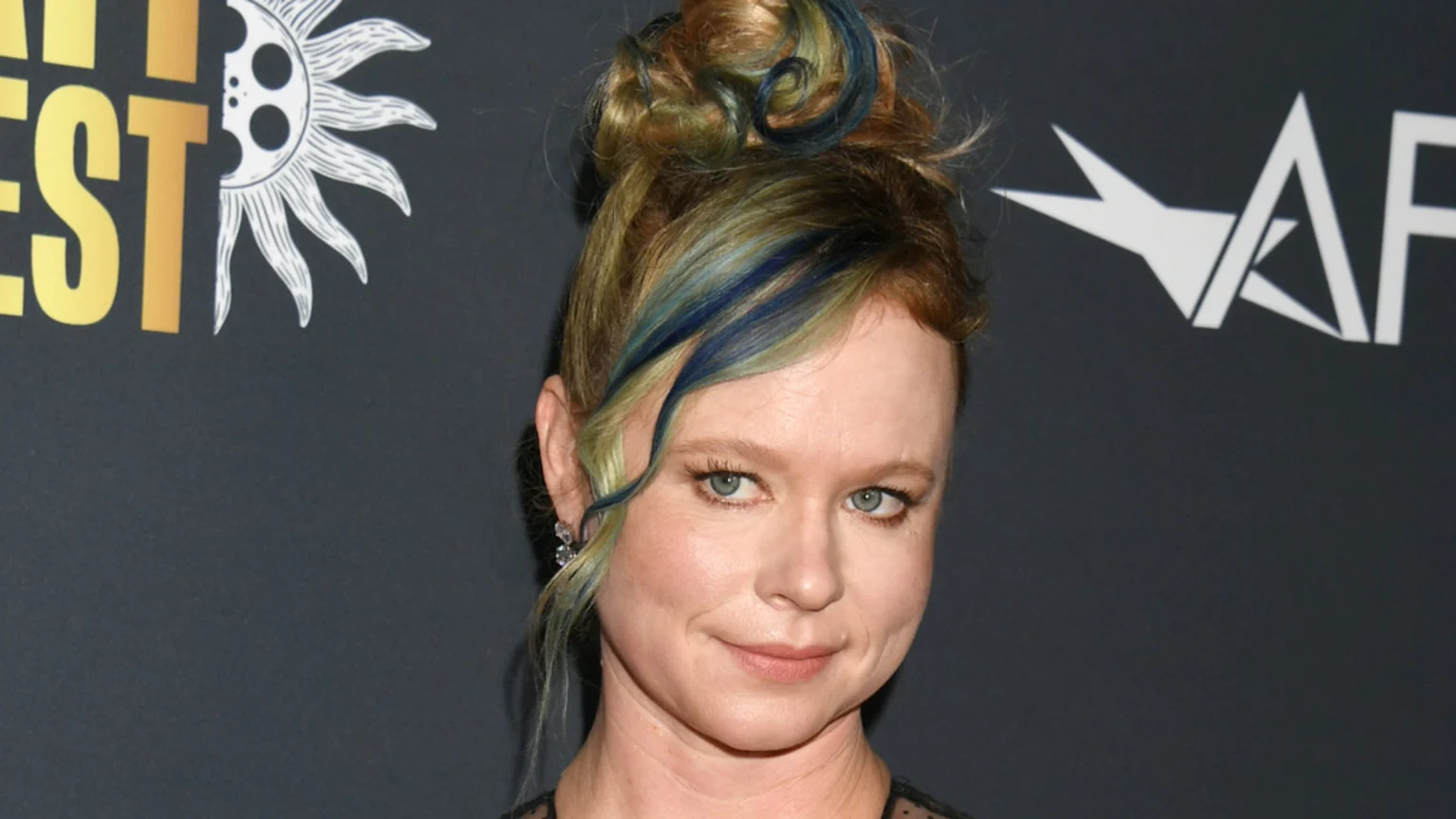 Close-up of actress Thora Birch smiling subtly, with reddish-brown hair in a messy bun featuring green and blue streaks, against a dark background with event logos.