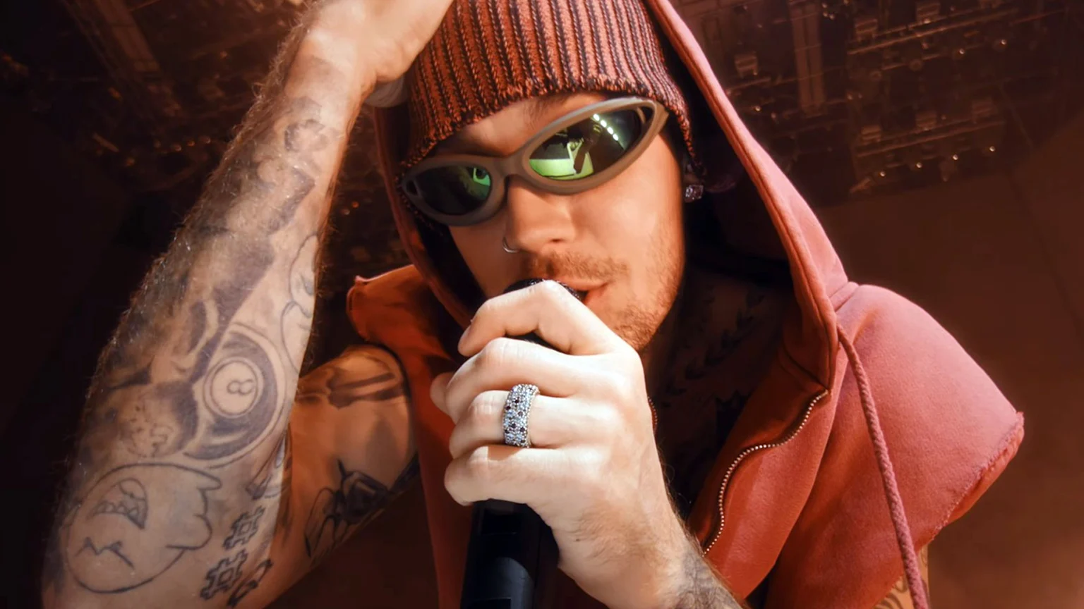 A man in a red beanie, hooded sweatshirt, and green-lensed sunglasses holds a microphone, showing tattooed arms and a multi-row studded ring.