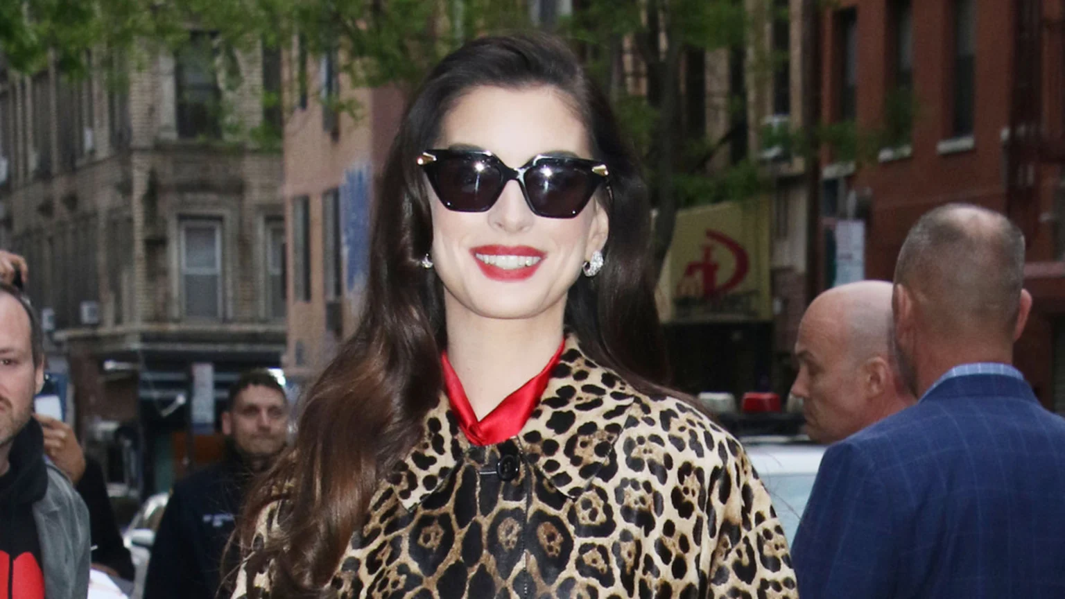 Anne Hathaway smiles, wearing black cat-eye sunglasses, red lipstick, and a leopard print coat over a red top, standing outdoors with blurred buildings and people in the background.