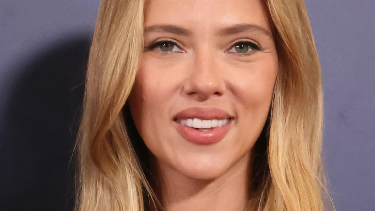 Close-up of a smiling woman with long blonde hair, green eyes, and light pink lipstick, against a plain dark blue background.