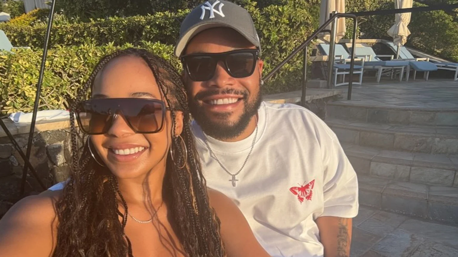 A close-up photo shows a man and a woman smiling and posing together outdoors in bright sunlight at a resort poolside. The woman wears oversized brown-tinted sunglasses, silver hoop earrings, and a delicate gemstone necklace. The man wears a grey baseball cap with a white 'NY' logo, dark rectangular sunglasses, a white t-shirt with a red butterfly logo, and a silver cross necklace. Lush green foliage, light blue cushioned lounge chairs, umbrellas, stone steps, and a black railing appear in the background.