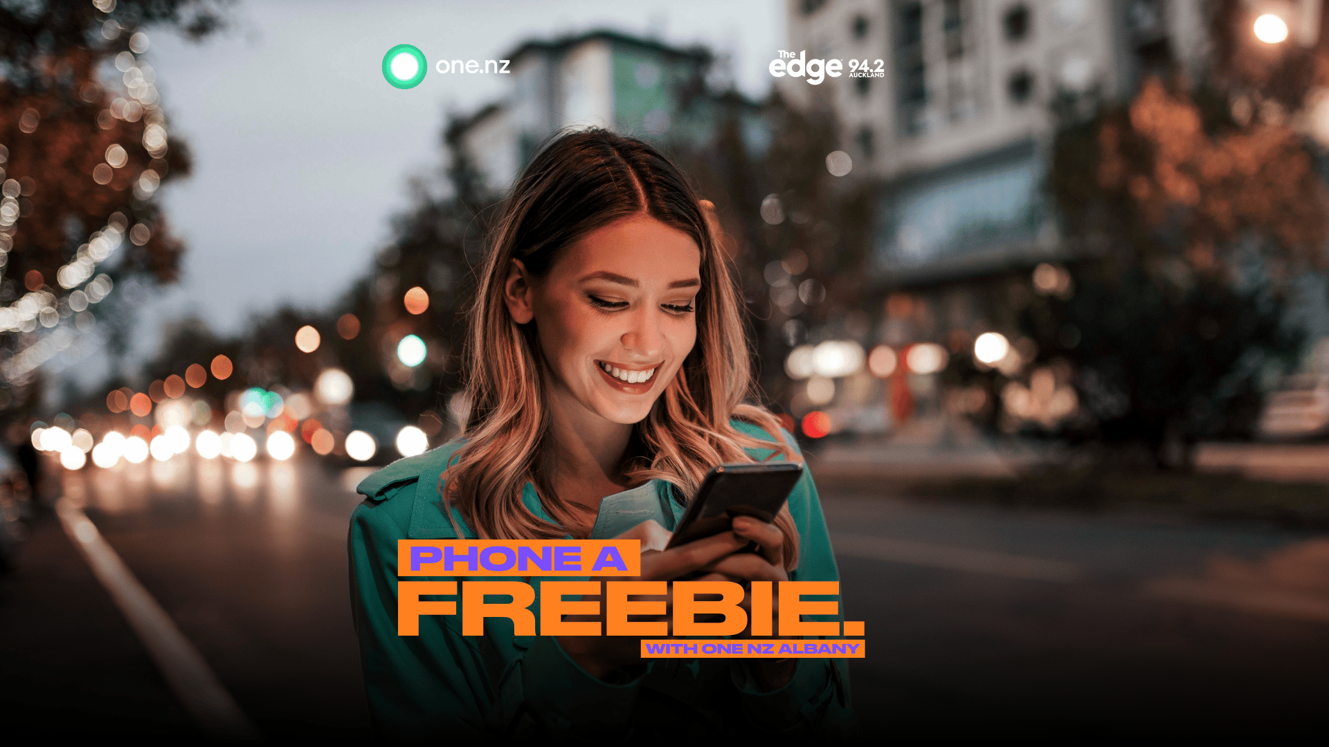 A smiling woman in a teal jacket looks at her phone on a blurred city street at night, with text "PHONE A FREEBIE. WITH ONE NZ ALBANY." and logos "one.nz" and "The Edge 94.2 Auckland".