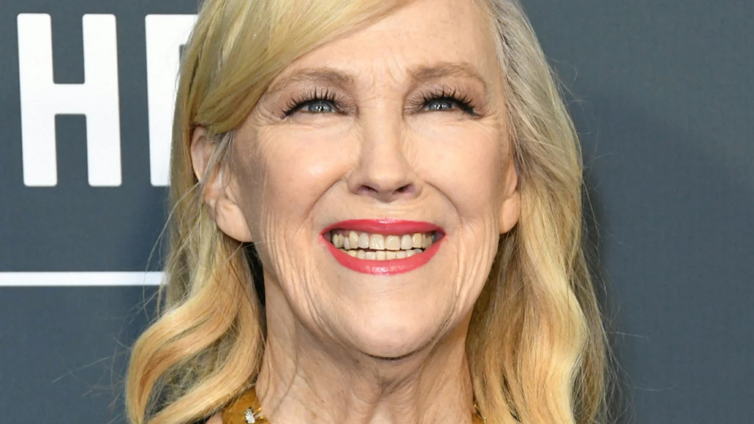 Close-up of a smiling older woman with blonde and grey hair, wearing red lipstick and mascara, against a dark background with white "HI" letters visible on the left.