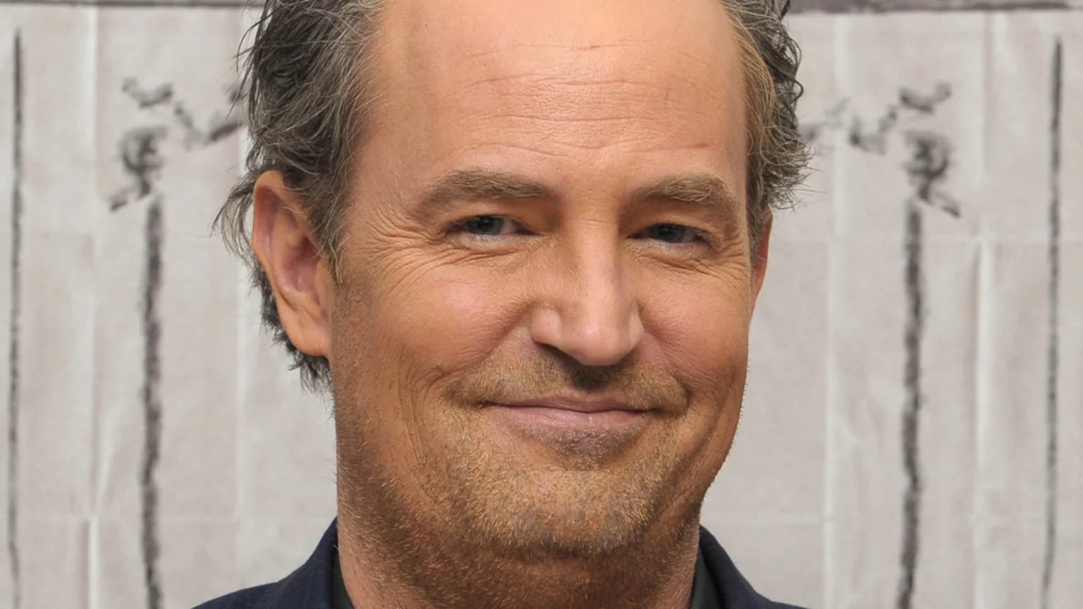 Close-up headshot of actor Matthew Perry smiling, with blue eyes, graying hair, and stubble, against a light background.