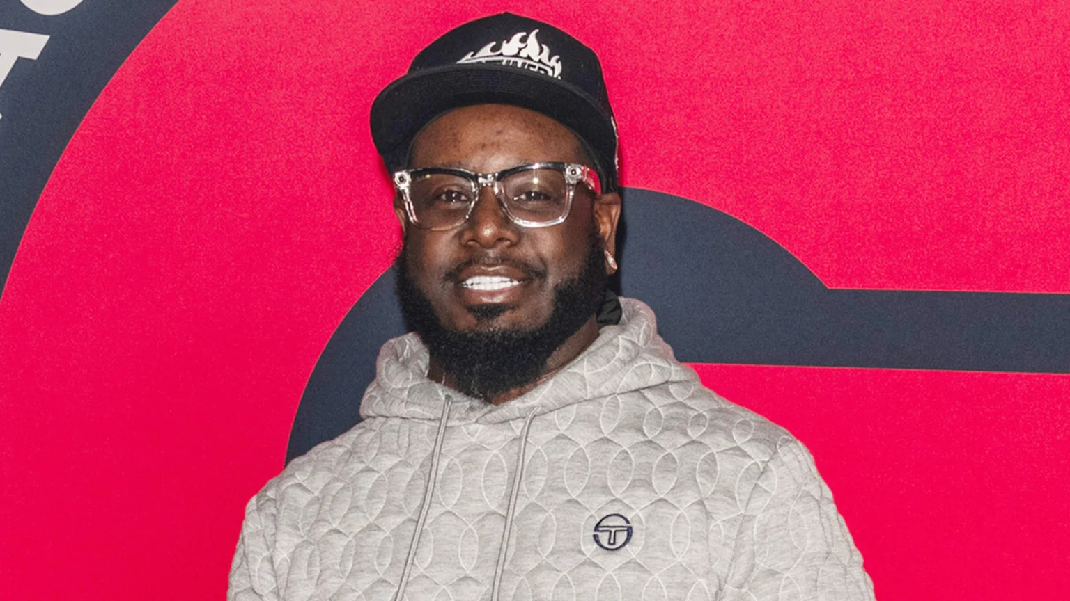A smiling Black man with a beard, clear glasses, and a black baseball cap wears a grey quilted hoodie, standing against a vibrant red background with dark blue graphic elements.
