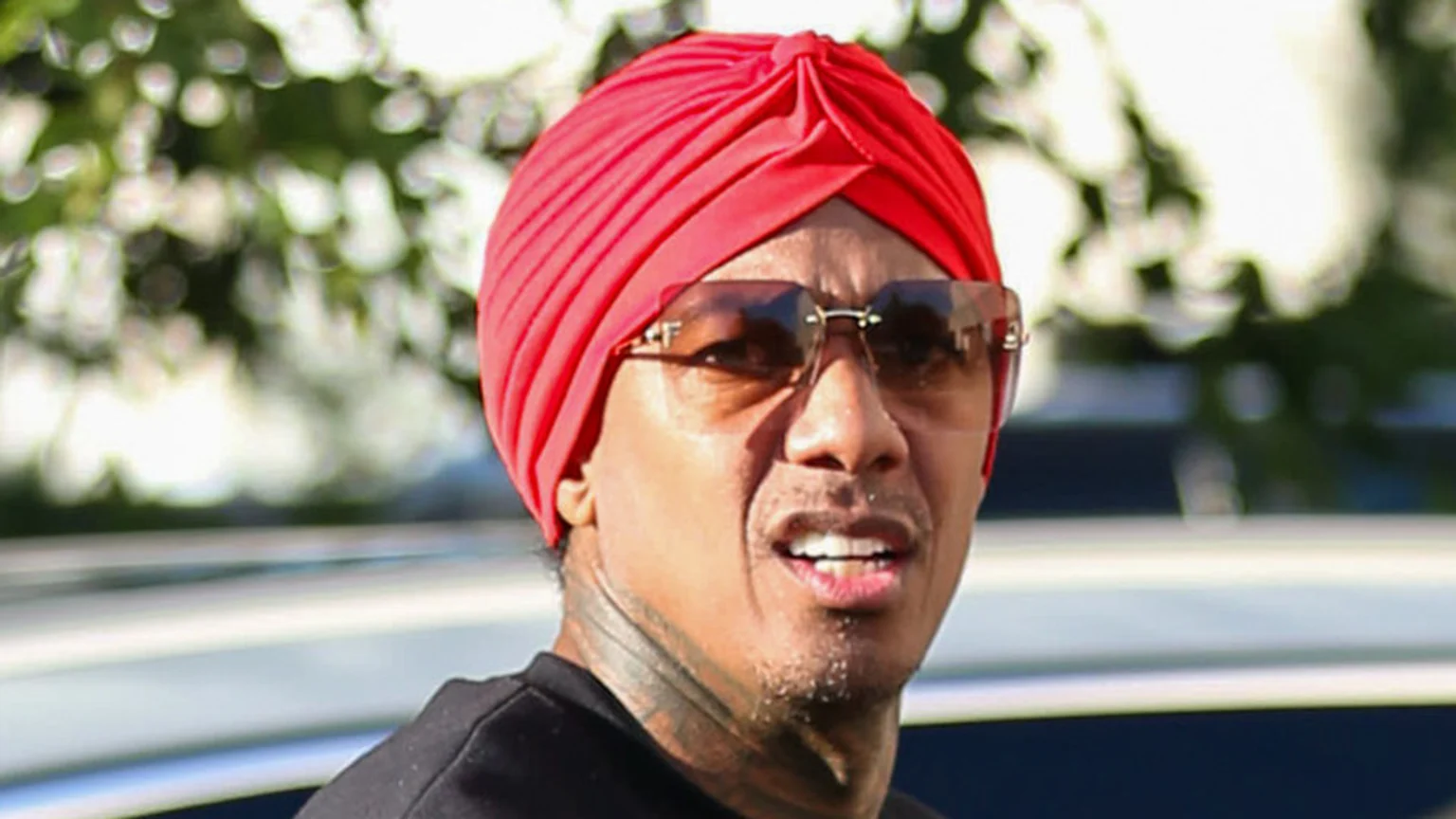 Nick Cannon wears a red turban and sunglasses, looking slightly to the side with a blurred green background.