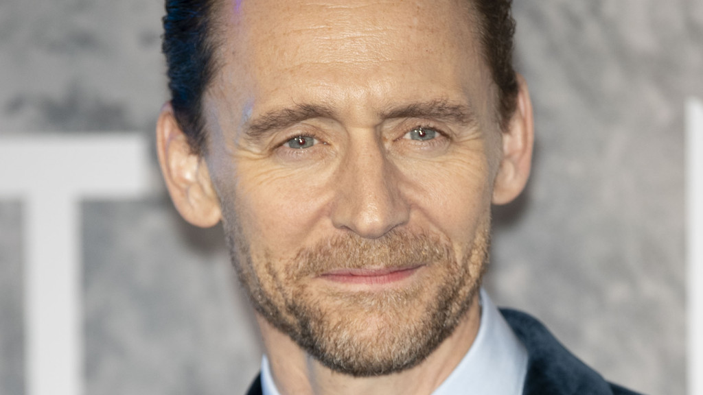 Close-up portrait of Tom Hiddleston, a light-skinned man with blue eyes, short brown hair, and a light brown beard, smiling slightly while looking directly forward, wearing a collared shirt and dark jacket.
