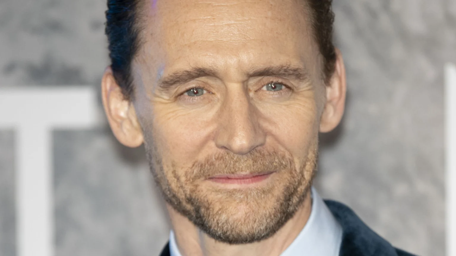 Close-up portrait of Tom Hiddleston, a light-skinned man with blue eyes, short brown hair, and a light brown beard, smiling slightly while looking directly forward, wearing a collared shirt and dark jacket.
