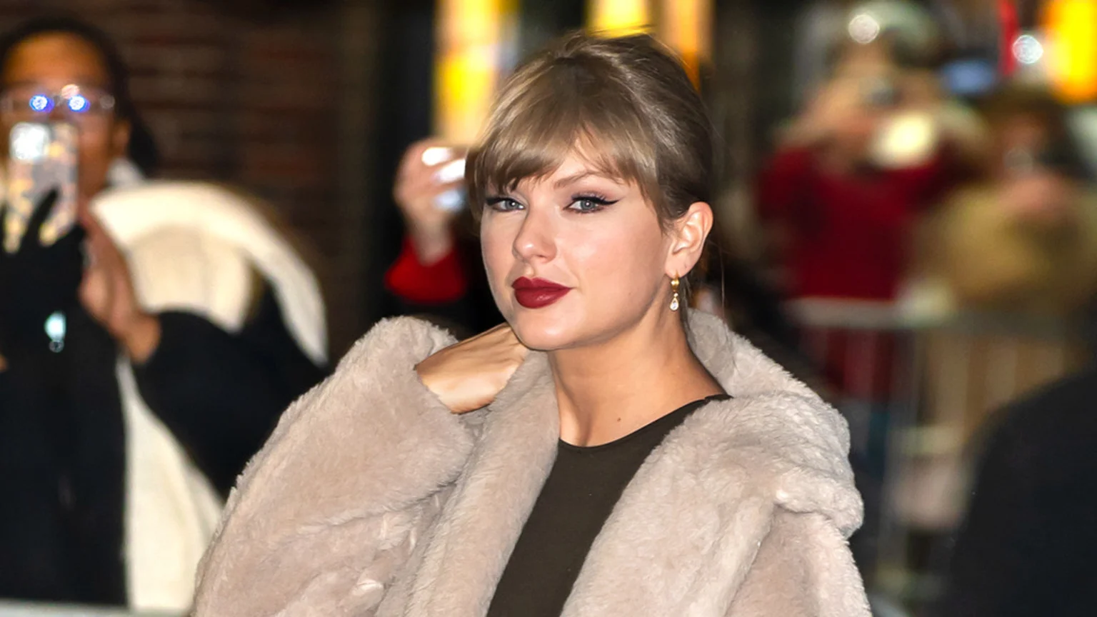 Taylor Swift, with blonde hair and red lipstick, wears a beige fluffy coat and looks at the camera, with blurred people and lights in the background.