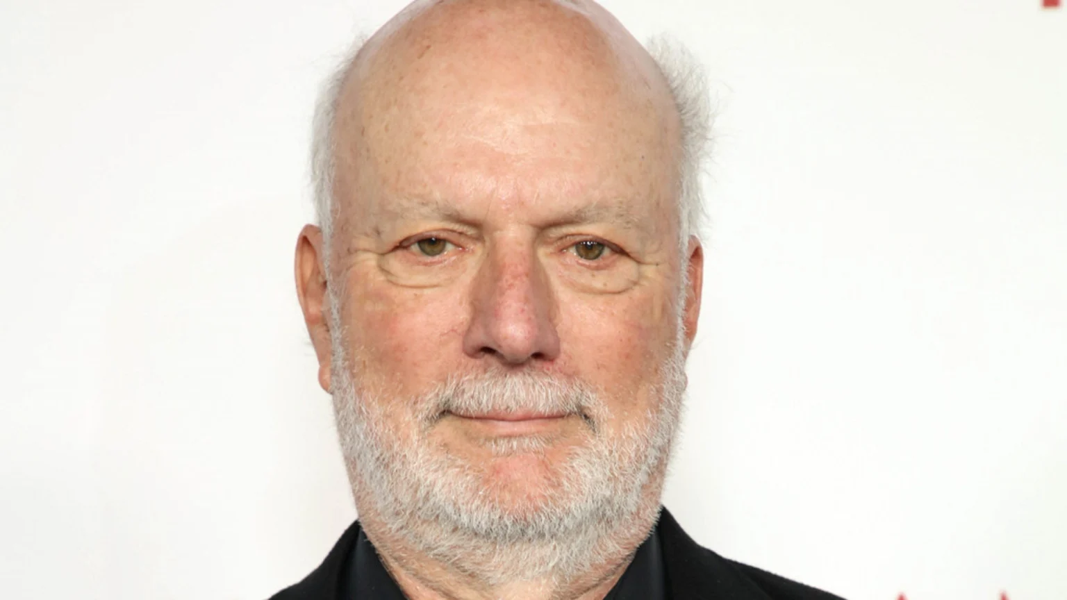 A close-up portrait of an older man with a bald head, white hair, and a white beard, looking at the camera with a slight smile.