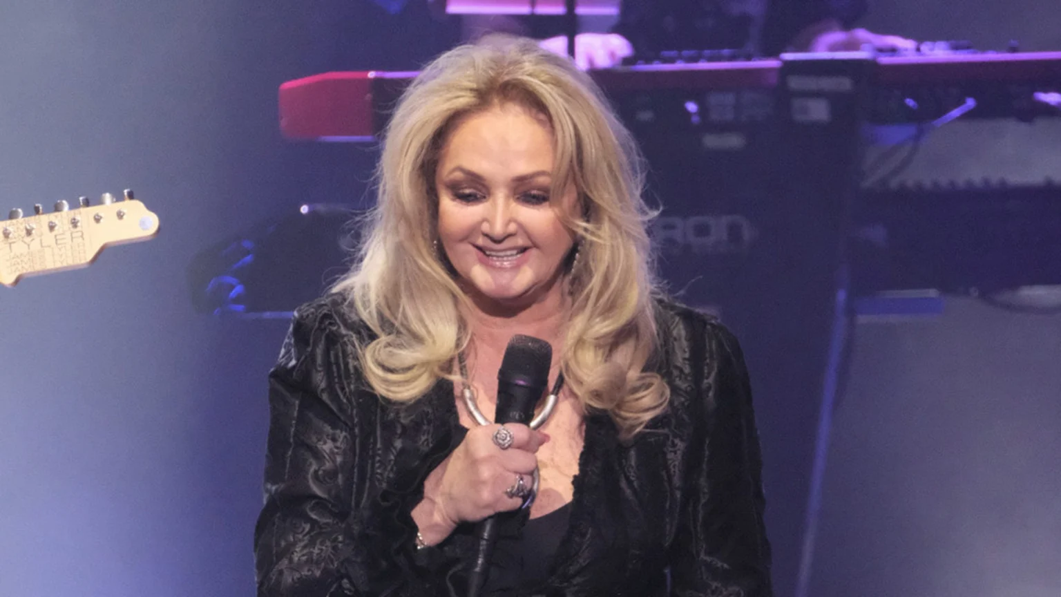 A smiling blonde woman with long hair in a dark, textured jacket holds a microphone, with blurry musical instruments visible in the stage background.