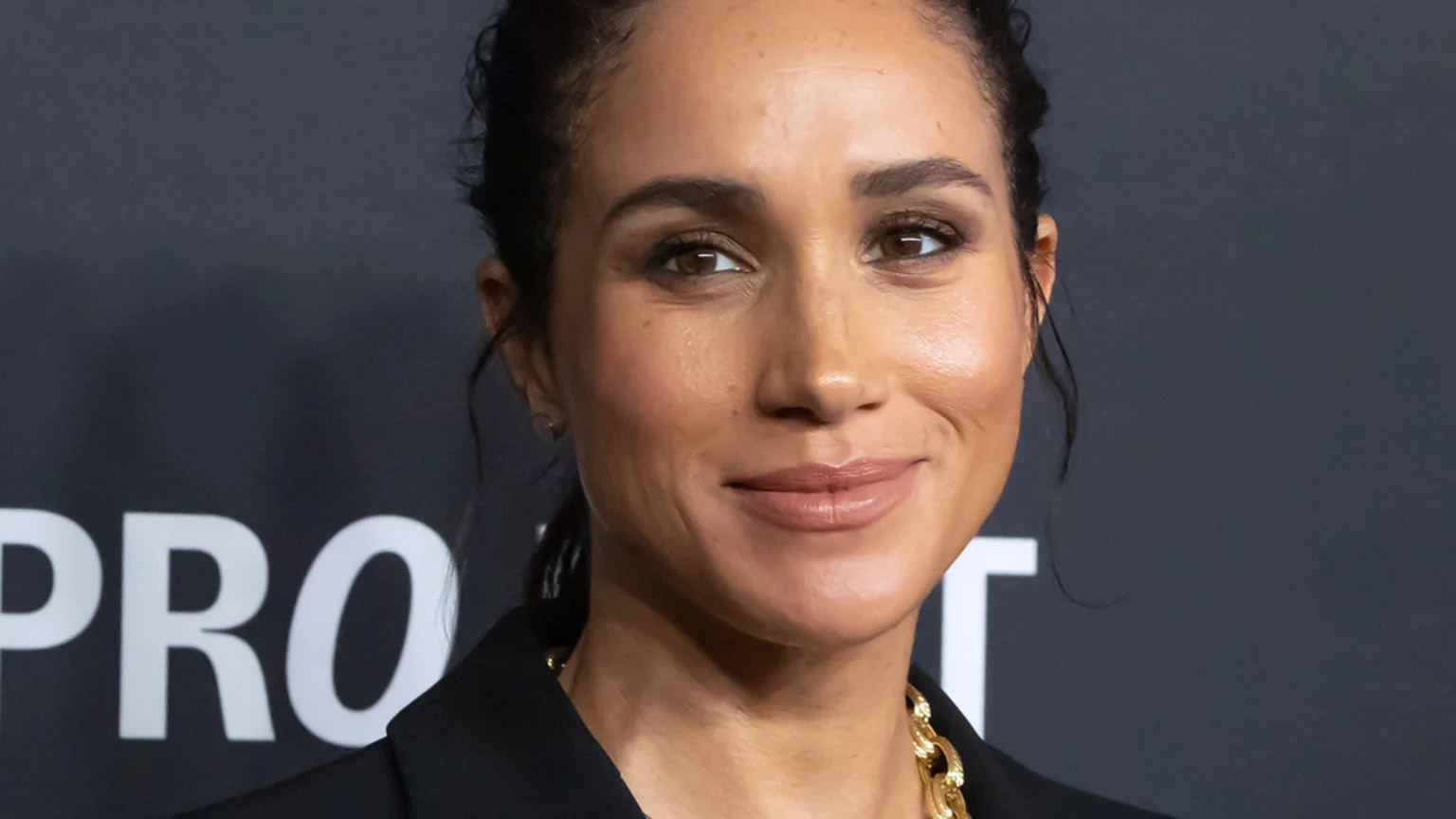 Meghan Markle, the Duchess of Sussex, smiles subtly, wearing a black top and a gold chain necklace, with her dark hair pulled back against a dark grey background.