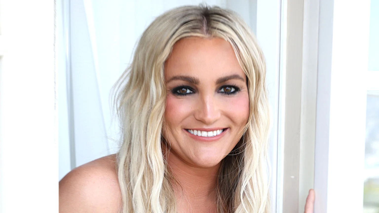 A smiling woman with long blonde hair, dark eye makeup, and a bare shoulder poses indoors against a bright, out-of-focus background.