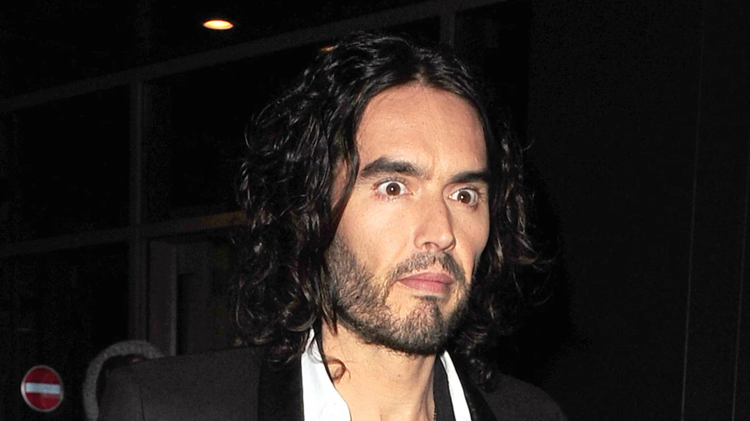 Russell Brand looks wide-eyed and surprised, with his dark curly hair and beard, wearing a dark jacket and white shirt, against a dark, blurry background with a "no entry" sign.