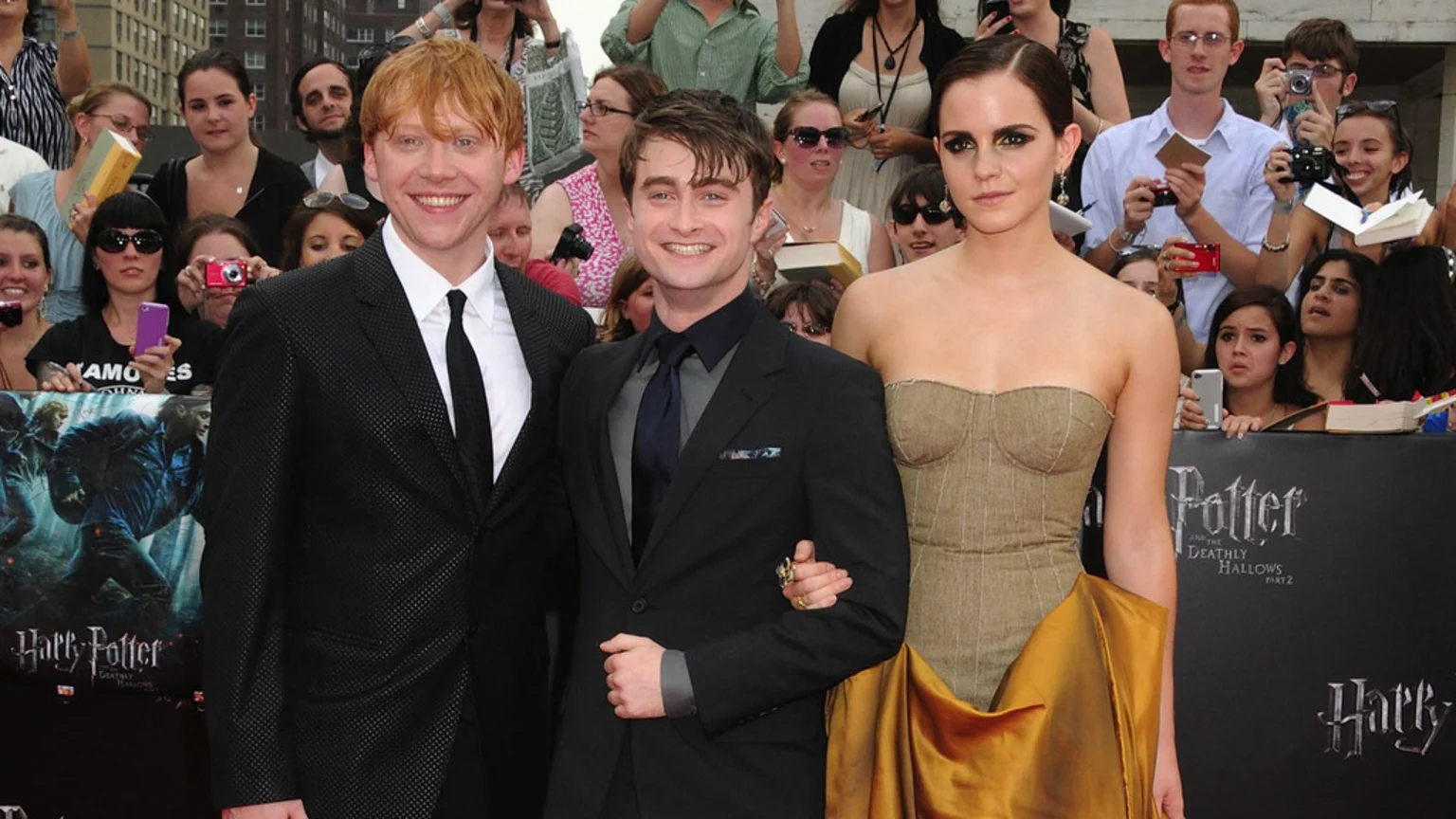 Rupert Grint, Daniel Radcliffe, and Emma Watson pose at a red carpet event, smiling in front of a Harry Potter banner and a crowd of fans.