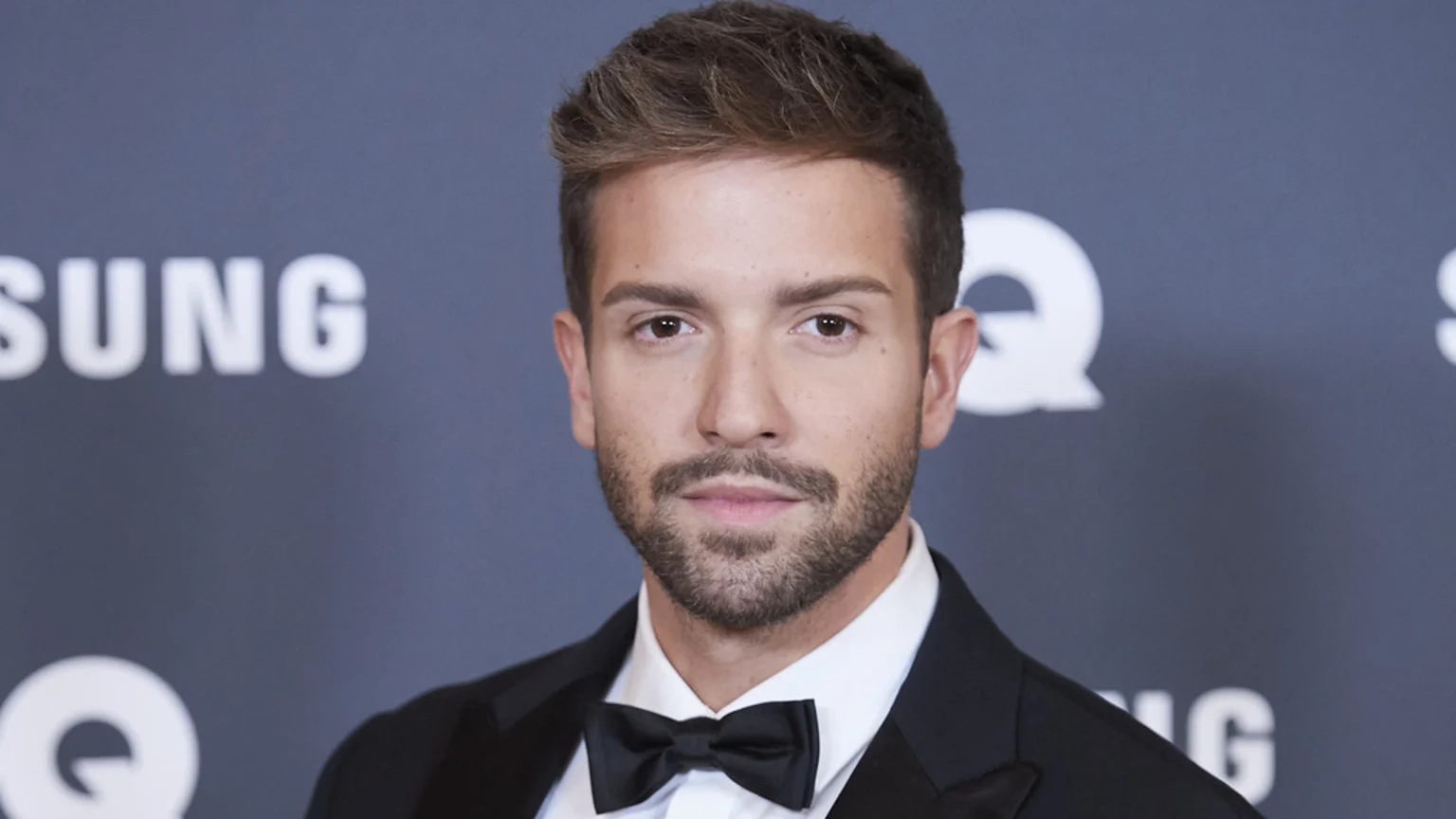 A man with brown hair and a beard, wearing a black tuxedo and bow tie, looks at the camera, with blurry "SAMSUNG" and "GQ" logos in the background.