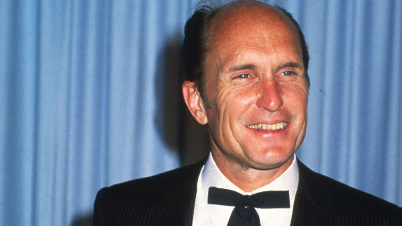 A brightly lit medium close-up portrait of actor Robert Duvall, smiling broadly with blue eyes, wearing a dark pinstripe suit jacket, white dress shirt, and black bow tie. He stands against a soft light blue fabric backdrop with vertical folds resembling drapes in a studio or event setting.