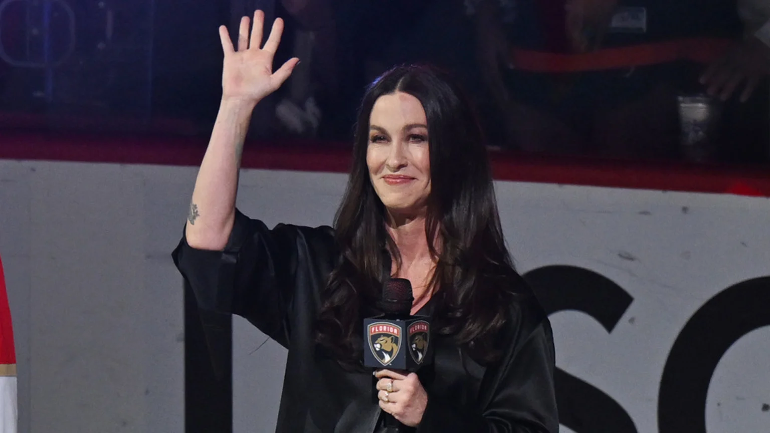 Alanis Morissette smiles and waves, holding a microphone with the Florida Panthers logo, against a dark, blurred background.