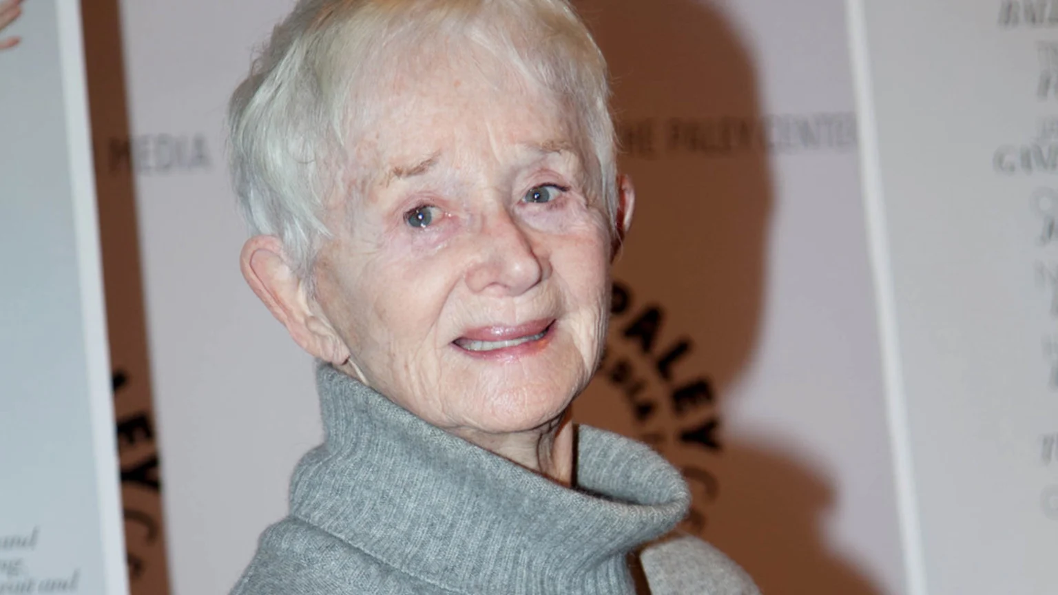 A portrait of an older woman, Barbara Barrie, with short white hair, blue eyes, and a slight smile, wearing a grey turtleneck sweater.
