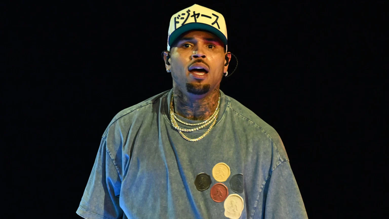 Chris Brown, with tattoos and a goatee, wears a cream and green cap, a faded blue t-shirt with circular patches, and multiple chains, looking directly with his mouth open against a dark background.